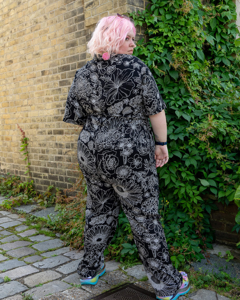 A model stands with their back turned, showcasing the black and white floral jumpsuit by Run & Fly. The jumpsuit features a bold, intricate flower print in white on a black background. The model, with pastel pink hair, pairs the ethical rayon jumpsuit with colourful platform shoes. The scene is set against a brick wall with greenery, promoting slow fashion and comfortable style.