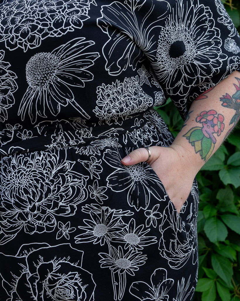 Close-up of the side of a black jumpsuit by Run & Fly featuring a detailed white floral print. The model’s hand rests in the jumpsuit’s pocket, showing off the relaxed fit and floral design. Tattoos on the model’s arm and leafy green background add contrast. Made from ethical rayon fabric, this jumpsuit is a statement piece for floral and alternative fashion lovers.