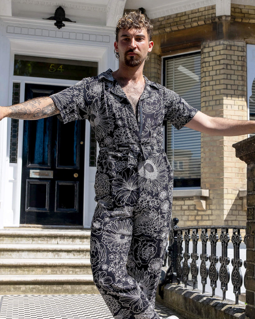 The Black and White Floral Rayon Jumpsuit worn by a masc non binary person with a brown curly mullet haircut and white trainers. They are stood in front of a doorway entrance with white ornate detailing, a black door, black ornate railing and black and white tiled steps. They are facing forward looking to camera with one leg crossed over the other and resting both arms on the railing. The black base jumpsuit features white floral outlines all over.