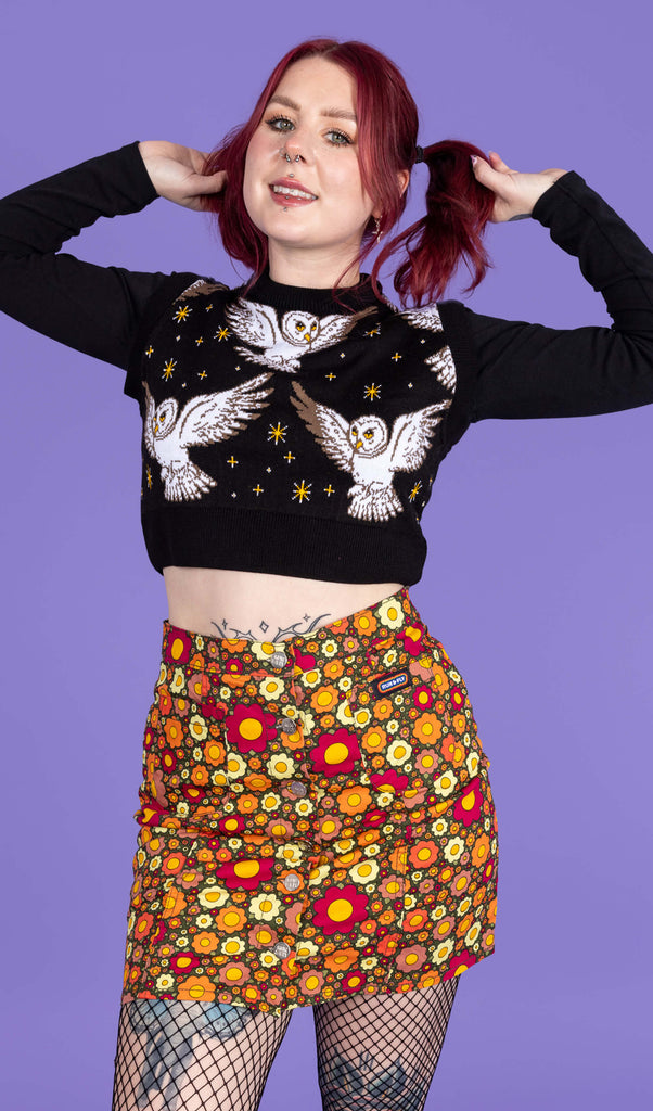 Florence is wearing Ditsy Floral A Line Skirt paired with a black jumper, fishnet tights and black boots. The skirt is a dark green colour and is covered in red, orange and yellow flowers. The model is posing with her hands in her hair and is smiling at the camera. The background of the photo is purple.