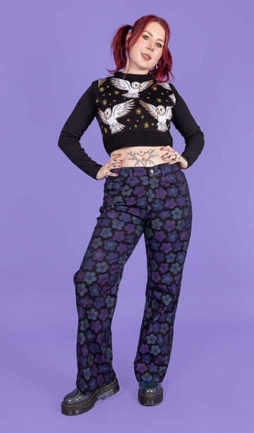 Florence a tattooed model with red hair is wearing DARK WASH 90's Black Magic Flower Wide Leg Stretch Jeans paired with a long sleeve black top, What a Hoot tank top and chunky boots. The black jeans have muted blue and purple flowers all over. The model is smiling at the camera with her hands on her hips and one leg turned outwards. The background of the photo is purple.