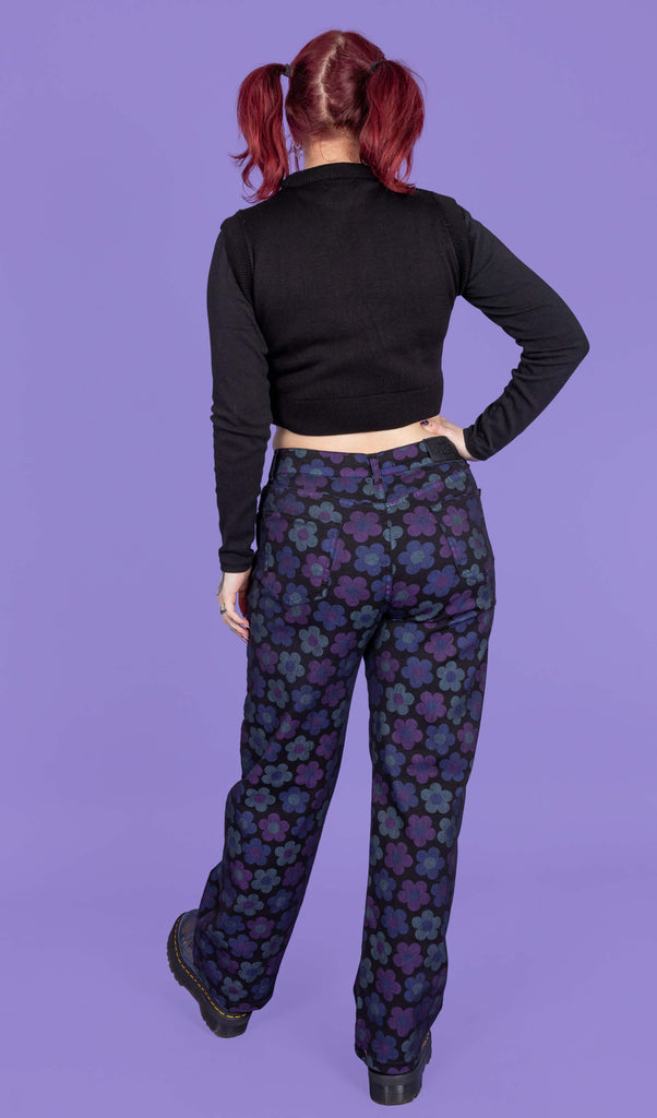 Florence a tattooed model with red hair is wearing DARK WASH 90's Black Magic Flower Wide Leg Stretch Jeans paired with a long sleeve black top, What a Hoot tank top and chunky boots. The black jeans have muted blue and purple flowers all over. Florence is stood with her back to the camera with one hand on her hip and one leg in front of the other. The background of the photo is purple.