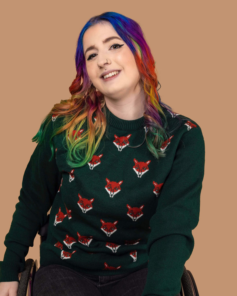 a white model with rainbow coloured hair is sat in their wheelchair in front of a beige backdrop with flowers on wearing Forest Green Fox Head Jumper paired with black jeans. The green long sleeved jumper has an all over print of fox heads on the torso. The model is smiling at the camera with one hand on wheelchair and other resting on their leg.