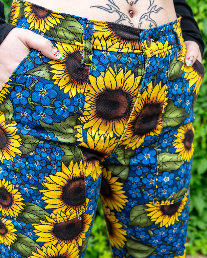 Close up of Run & Fly x Jen James Design Forget Me Not Charity High Waist Straight Leg Jeans. The print is an all over bright yellow sunflower and blue forget me not floral design. The model's hands are resting in the pockets of the jeans and they are paired with a black crop top.
