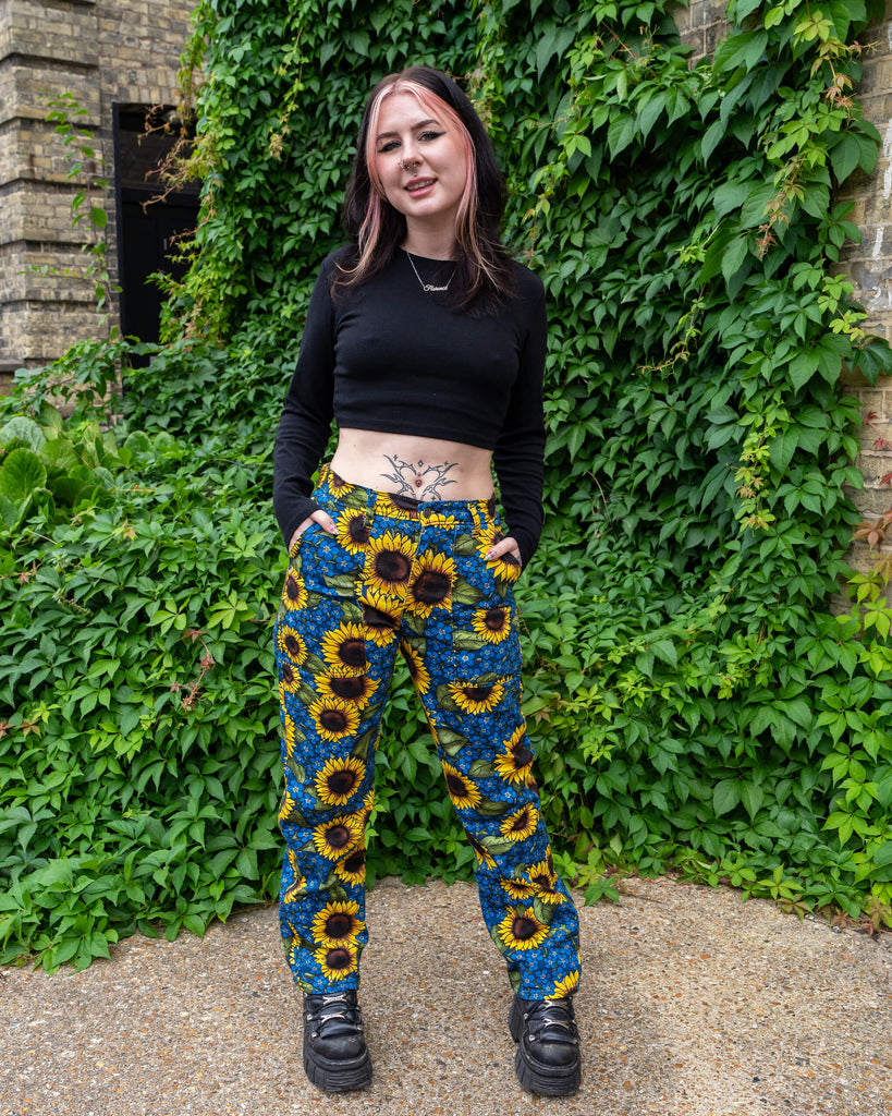 Florence is stood in front of greenery wearing Run & Fly x Jen James Design Forget Me Not Charity High Waist Straight Leg Jeans paired with a black crop top and chunky boots. The print is an all over bright yellow sunflower and blue forget me not floral design. She has pink and black hair and is facing the camera posing with her hands in he jean pockets 

