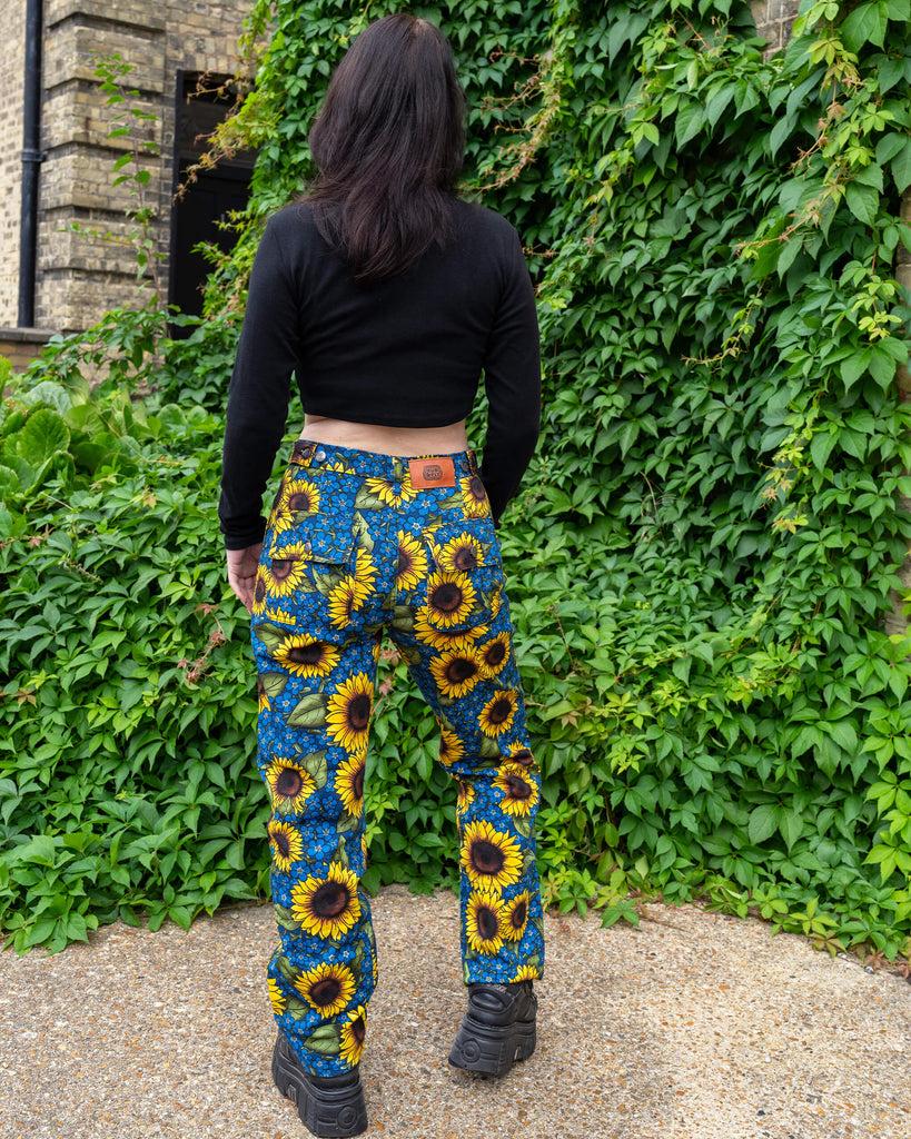 lorence is stood in front of greenery wearing Run & Fly x Jen James Design Forget Me Not Charity High Waist Straight Leg Jeans paired with a black crop top and chunky boots. The print is an all over bright yellow sunflower and blue forget me not floral design. She is stood with her back to the camera with er arms resting her sides.