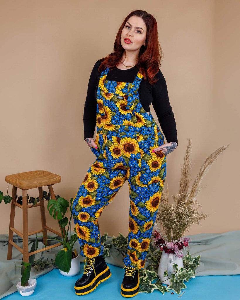 Aemelia Fox is stood in a photography studio in Hove with a tan colour backdrop wearing Run & Fly x Jen James Design Forget Me Not Charity Stretch Twill Dungarees with a long sleeve black top underneath and boots. The dungarees have an all over blue forget me not and yellow sunflower print. Aemelia is posing at the camera with one leg bent and her hands in the dungaree pockets.