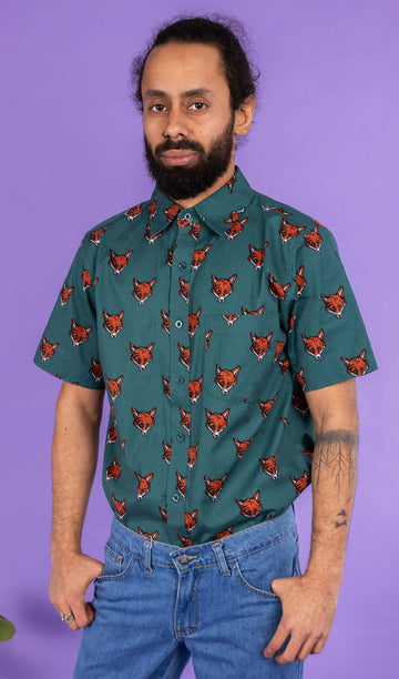 Richard, a hispanic male model with dark hair in a bun and a beard, is stood in a photography studio in Hove in front of a lilac backdrop wearing Preppy Foxy Short Sleeve Shirt with blue jeans. The emerald green shirt has an all over print of fox heads. Richard is posing facing the camera with his thumbs resting in the front pockets of his jeans. The photo is cropped at the thighs.