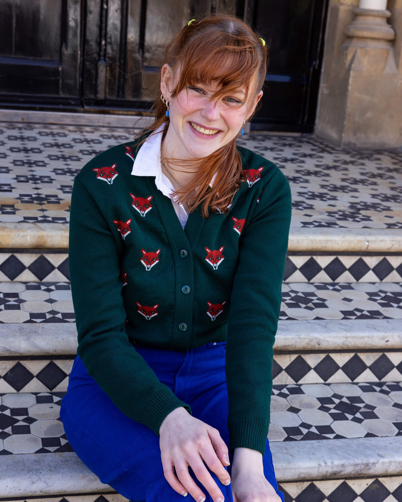A model sitting on tiled steps wearing a green cardigan with red fox head designs and vibrant blue trousers. The model smiles warmly, with ginger hair tied up and a white collared shirt beneath the cardigan. Ethical slow fashion by Run & Fly.