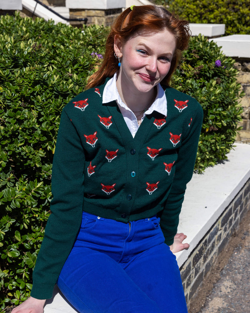 A model sitting outdoors in a green cardigan featuring red fox head designs and vibrant blue trousers. The model smiles softly, seated on a low wall surrounded by green shrubbery. Ethical slow fashion by Run & Fly, promoting playful knitwear.
