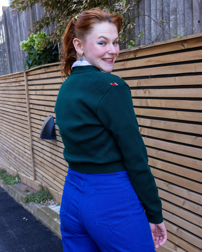 A model looking over their shoulder, wearing a forest green cardigan with red fox head designs and blue trousers. The model is smiling while standing outdoors, posing in front of a wooden fence. Ethical slow fashion by Run & Fly.