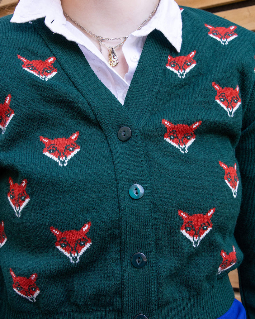 Close-up of a forest green cardigan featuring repeated red fox head designs. The cardigan has black buttons and is worn over a white collared shirt. Ethical slow fashion by Run & Fly, showcasing intricate knitwear details with bold animal motifs.