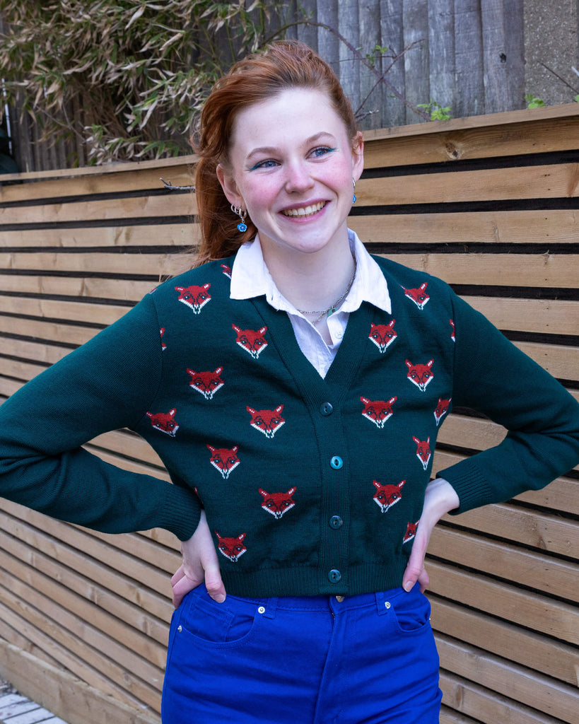 A model standing outdoors, hands on hips, wearing a forest green cardigan with red fox head designs and vibrant blue trousers. The model is smiling brightly, with ginger hair pulled back in a ponytail, and is posing in front of a wooden fence. Ethical slow fashion by Run & Fly.