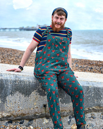 A ginger bearded model with a large septum ring, blue beanie, rainbow stripe navy top and black boots wearing the Green Fox Stretch Corduroy Dungarees on brighton beach with the pier in the background. The green Fox Stretch Corduroy Dungarees are a dark forest green with bright orange traditional fox heads all over.