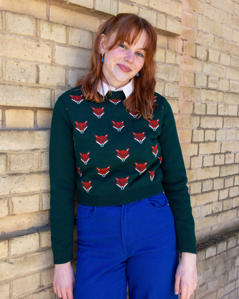  Model is wearing Green Fox Cropped Jumper, paired with a white collared shirt and bright blue jeans. The jumper is a dark forest green colour with a print of multiple red foxes on the front torso. Model is leant against a wall, smiling at camera.