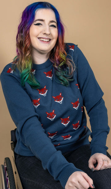 Eliza with rainbow hair is sat in their wheelchair wearing the blue fox head jumper with black stone wash flares. They are leaning forward with both hands resting on their lap and smiling to camera. The blue fox head jumper is a petrol blue base colour with red and white fox heads repeating over the middle of the jumper segment, the arms and back are plain petrol blue.