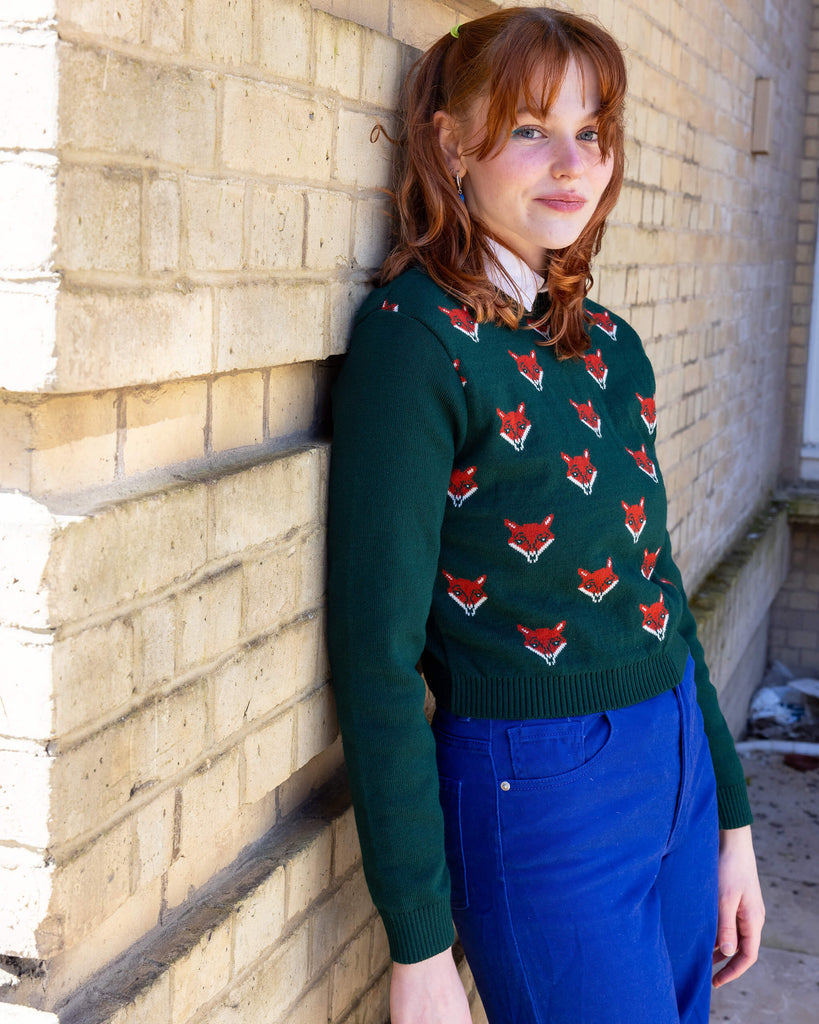 Model is wearing Green Fox Cropped Jumper, paired with a white collared shirt and bright blue jeans. The jumper is a dark forest green colour with a print of multiple red foxes on the front torso. Model is leant against a wall, smiling at camera.