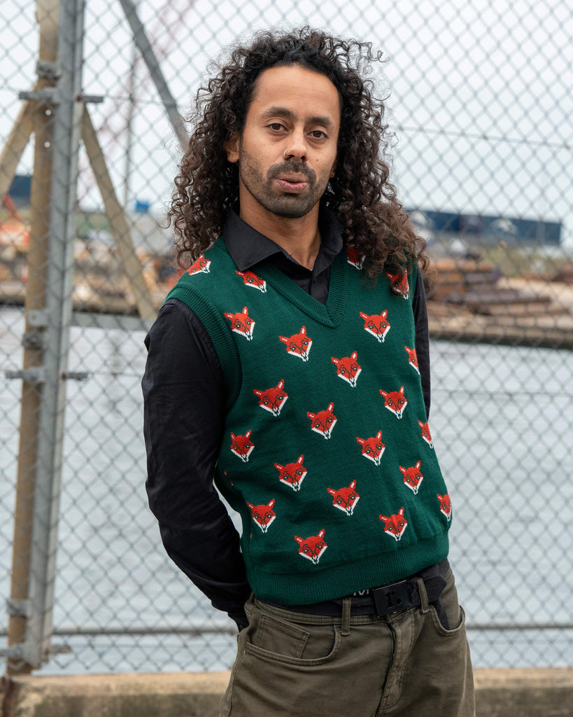 Model Rich wears the Run & Fly Fox Head Tank Vest featuring red fox faces on a green knit. Styled over a black shirt for a fitted unisex look.