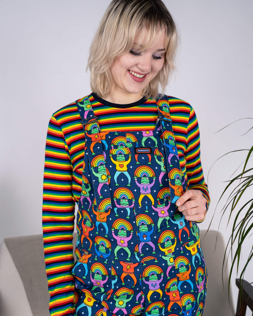 Smiling model looking down while wearing Run & Fly Rainbow Frogs Stretch Twill Dungarees and a rainbow-striped top. The vibrant frog print adds whimsy to ethical slow fashion.