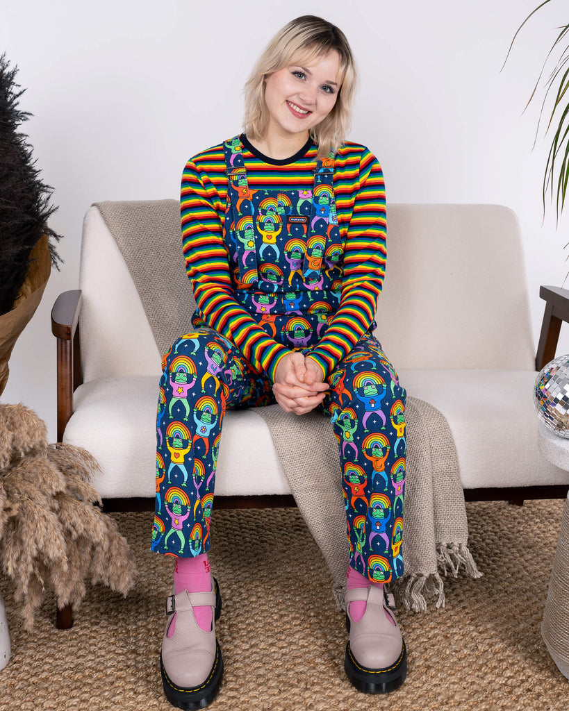 Model seated with hands clasped, showcasing Run & Fly Rainbow Frogs Stretch Twill Dungarees paired with a cheerful rainbow-striped top. Ethical slow fashion with playful charm.