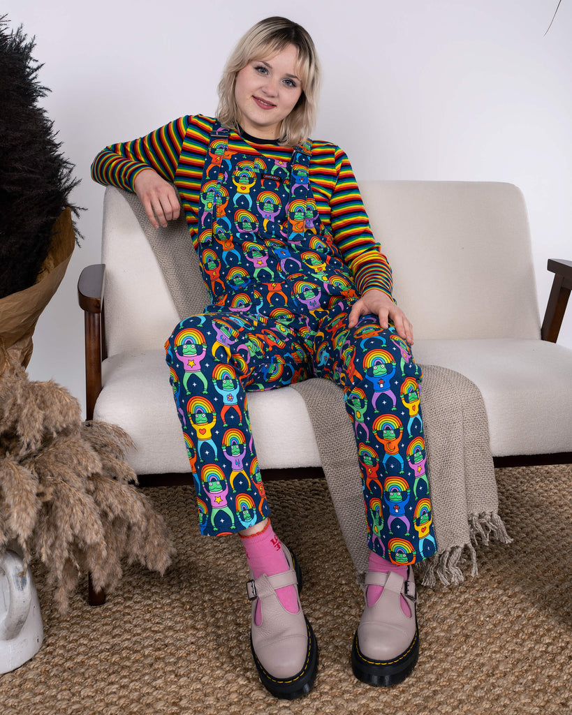 Model sitting on a cream sofa, wearing Run & Fly Rainbow Frogs Stretch Twill Dungarees styled with a rainbow-striped top. The frog and rainbow design makes a vibrant statement in ethical fashion.