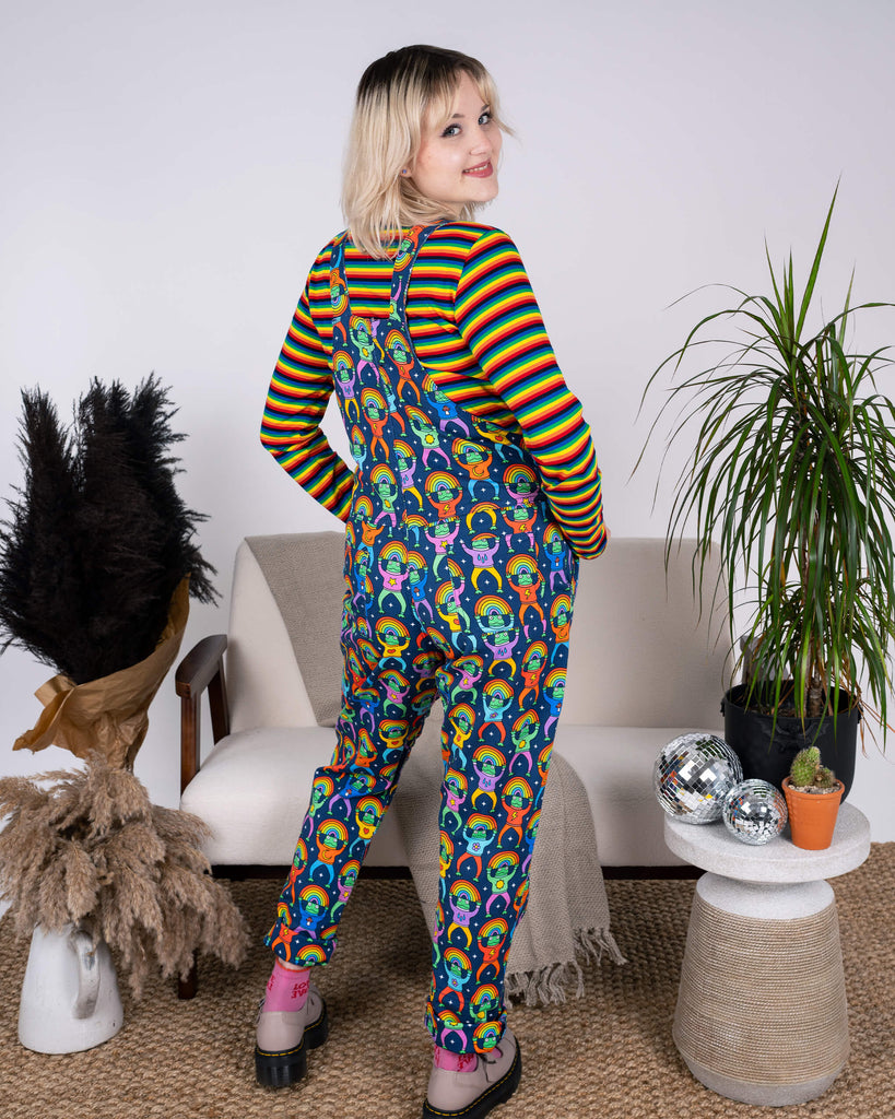 Back view of a model wearing Run & Fly Rainbow Frogs Stretch Twill Dungarees, paired with a rainbow-striped top, standing in a cosy, plant-filled setting. Ethical and fun slow fashion.