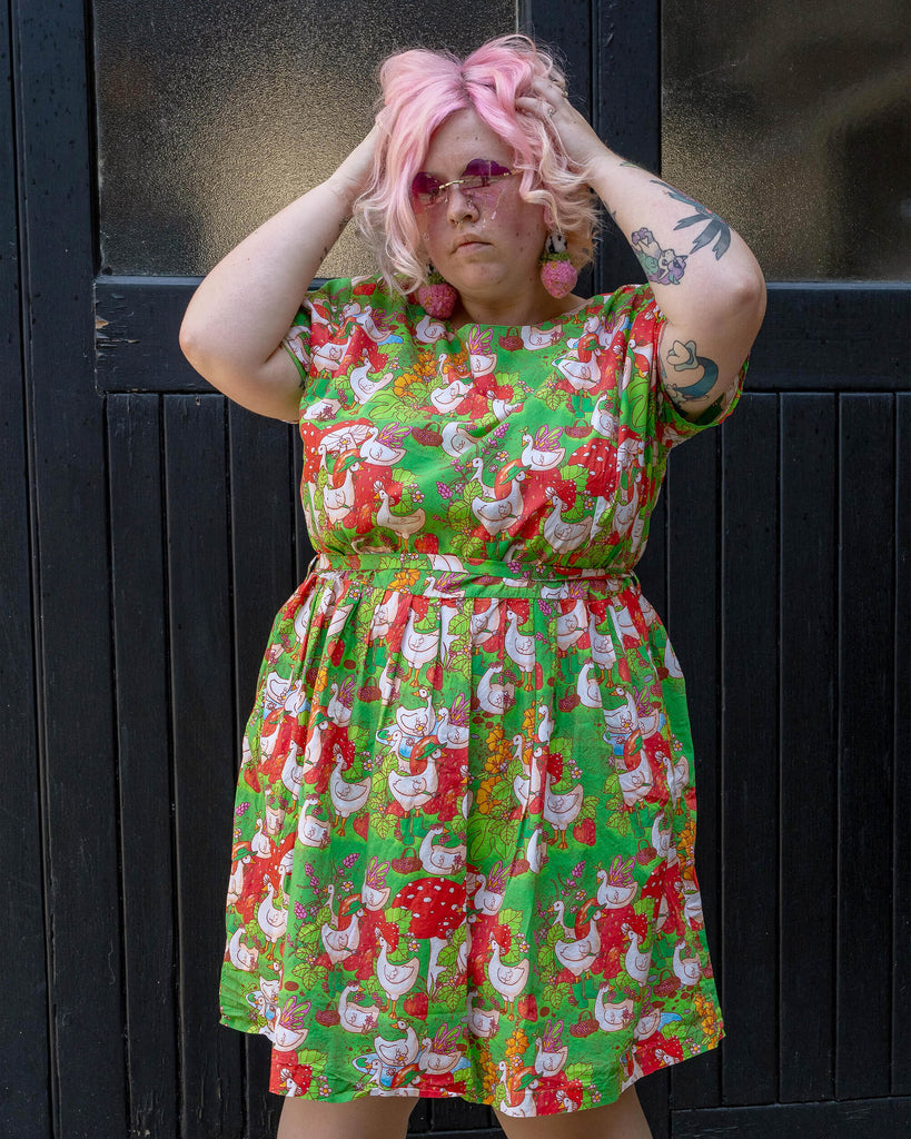 A plus-size model with pastel pink hair and pink sunglasses showcases a colourful dress with a playful geese and garden motif. The dress, from the ethical brand Run & Fly, features a fitted waist and short sleeves, set against a dark background.