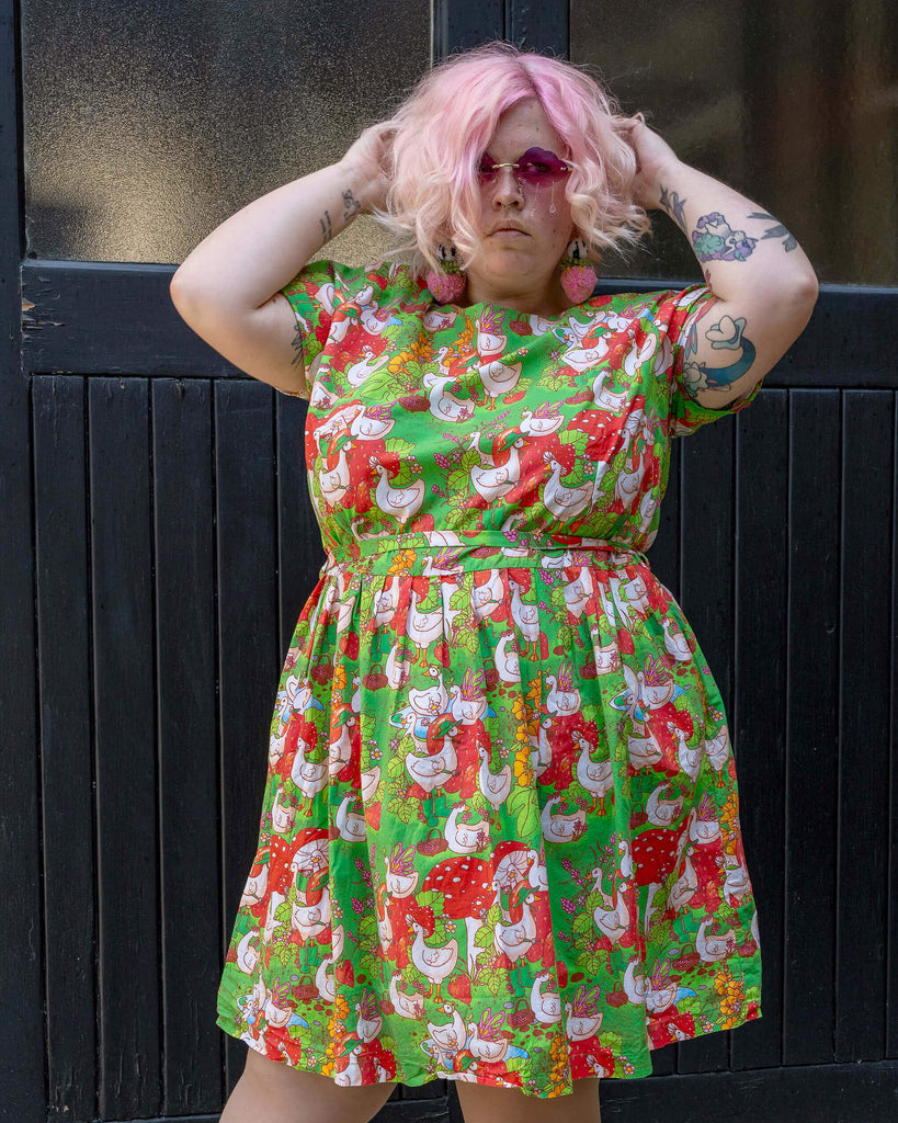 A model with pink hair and pink sunglasses poses confidently, wearing a vibrant green dress adorned with a whimsical geese and garden print. The dress is from Run & Fly, a small ethical slow fashion brand. They stand in front of a dark wooden door.