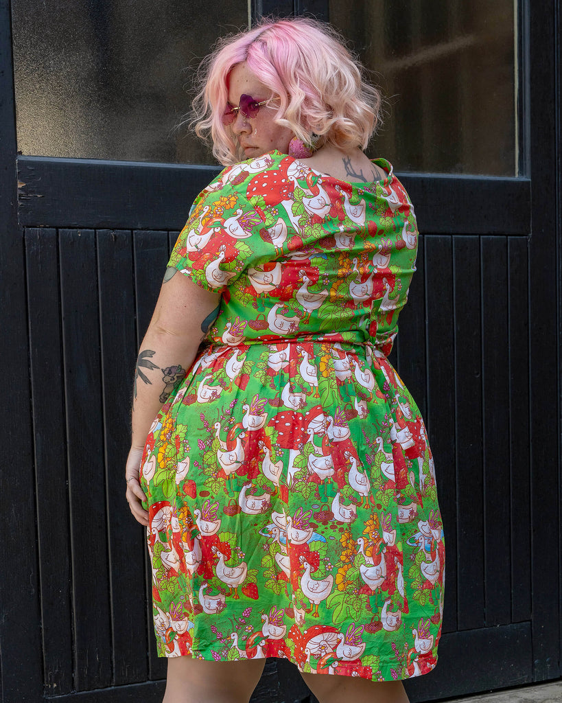 A plus-size model with pastel pink hair is seen from the back, showcasing the detailed geese and garden print on their green dress. The Run & Fly dress is part of the brand's ethical slow fashion line, providing a playful yet responsible fashion choice.