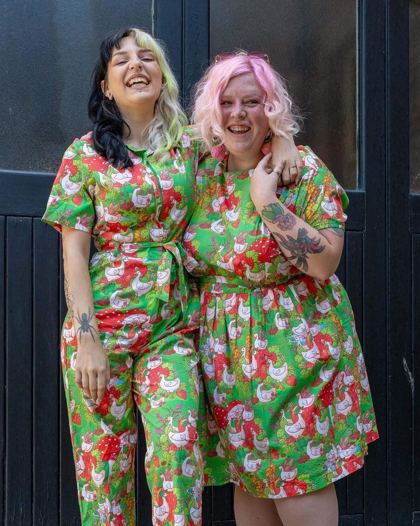 Two models one with black and blonde hair and the other with pink hair, laugh and pose together. They are wearing matching green outfits with a geese and garden print by Run & Fly, showcasing the brand's fun and ethical slow fashion designs.