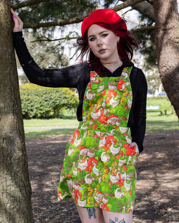 The Run & Fly x The Mushroom Babes In The Geese Garden Stretch Twill Pinafore Dress being worn by Flo outside in a park with a long sleeve black top, black trainers and red mushroom beret. She has red and black hair and tattoos, she is facing forward smising leaning one hand on a tree with the other in the dress pocket. The print features geese wearing various cute hats, such as mushrooms and frogs, collecting flowers and strawberries on green grass.