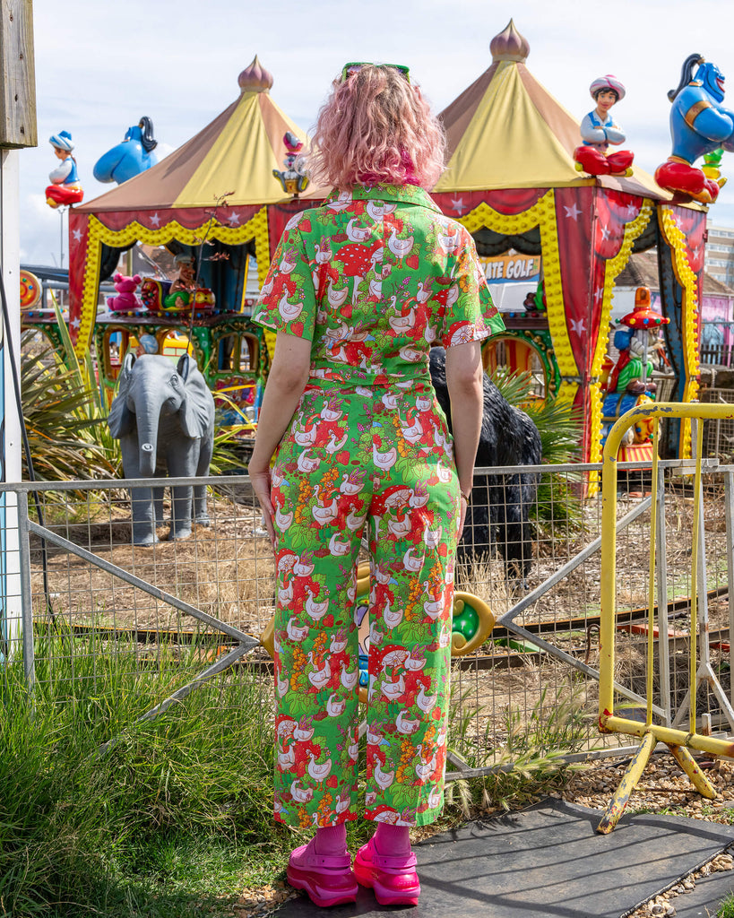 A person with curly pink hair stands with their back facing the camera, wearing a bright green jumpsuit covered in whimsical geese, strawberry, and mushroom illustrations. The outfit is cinched at the waist with a matching belt. The model is in front of a colourful fairground scene. Run & Fly ethical slow fashion brand.
