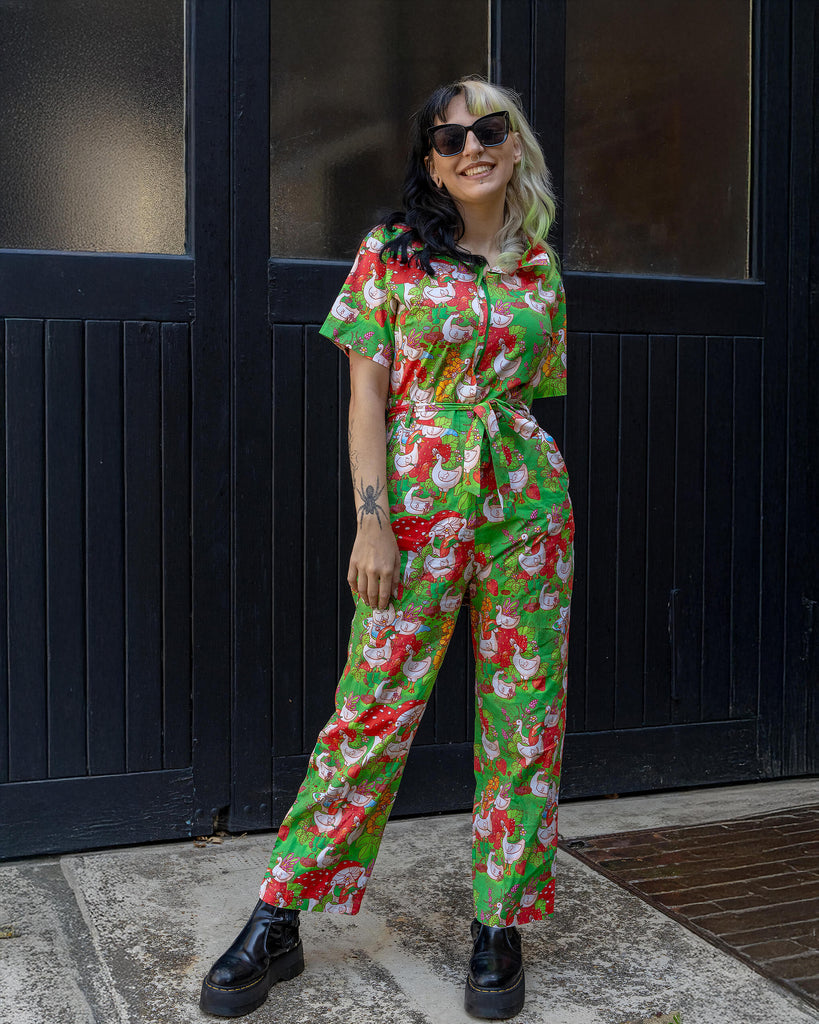 A model with two-tone black and blonde hair poses in a colourful green jumpsuit by Run & Fly. The whimsical print features geese, strawberries, and mushrooms. Ethical and slow fashion with a fun, bold style. The model wears chunky black boots.