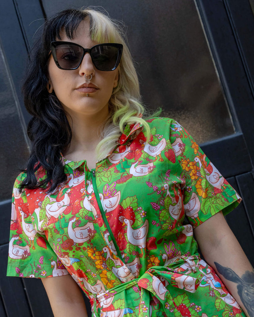 A close-up shot of a model with two-tone black and blonde hair, wearing sunglasses and a green jumpsuit by Run & Fly. The outfit features a quirky geese and strawberry print, reflecting the brand’s playful approach to ethical slow fashion.