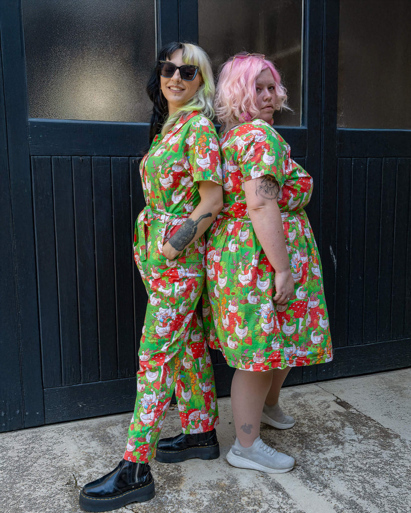 Two models stand back-to-back wearing matching green Run & Fly outfits, one in a jumpsuit, the other in a dress. Both pieces showcase a whimsical geese and strawberry print. Run & Fly’s ethical slow fashion ethos shines through in this fun and bold design.
