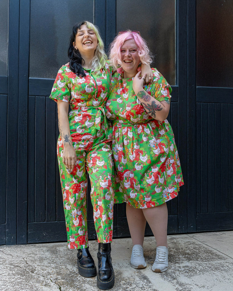 Two smiling models wearing matching green outfits from Run & Fly, one in a jumpsuit and the other in a dress. The playful print features geese, strawberries, and mushrooms. The brand promotes ethical slow fashion with fun, bold designs. One model has two-tone hair and the other has pink curls.