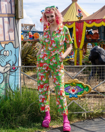 A model with pink curly hair smiles, wearing a vibrant green jumpsuit covered in geese and strawberries. The jumpsuit has short sleeves, a collar, and a belted waist. The colourful fairground background complements the fun print. Run & Fly, small ethical slow fashion company.