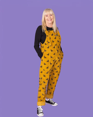 A three-quarter view of an elderly woman with long blonde hair wearing a mustard yellow corduroy dungaree with black bee patterns over a black long-sleeve shirt. She has her hands in the front pockets of the dungarees and is smiling. The background is a solid purple.