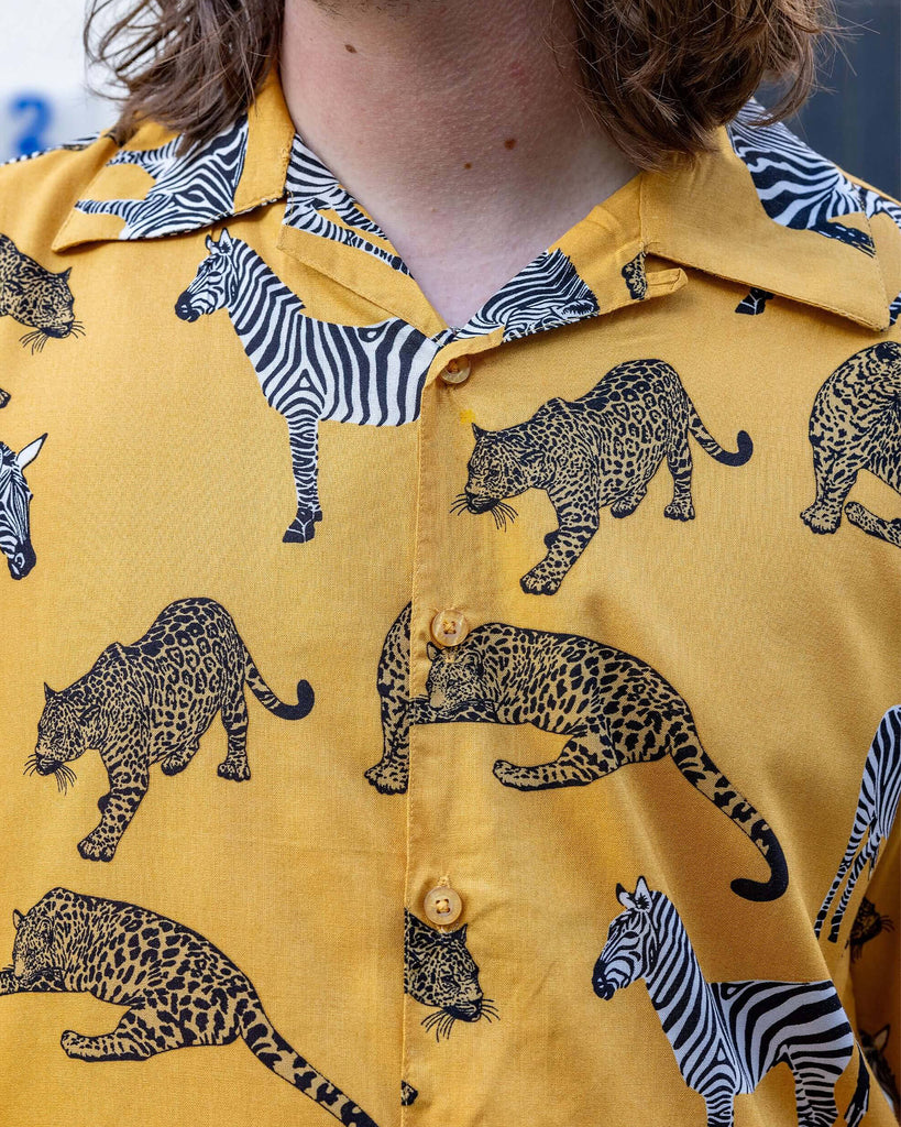 close up of white male model wearing Golden Zebra Short Sleeve Rayon Shirt. The shirt is a yellow gold colour with an all over print of zebras and big cats. The close up shows the print and yellow buttons on the shirt. 