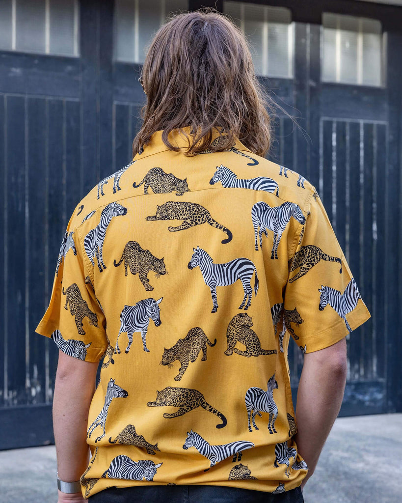 white male model with long hair and glasses wearing Golden Zebra Short Sleeve Rayon Shirt paired with black jeans. The shirt is a yellow gold colour with an all over print of zebras and big cats. The model is stood outside in Hove posing with his back to the camera to show the back of the shirt.
