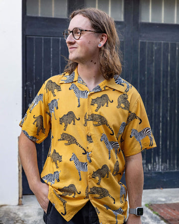 white male model with long hair and glasses wearing Golden Zebra Short Sleeve Rayon Shirt paired with black jeans. The shirt is a yellow gold colour with an all over print of zebras and big cats. The model is stood outside in Hove posing toward the camera with one hand in his jeans pocket.