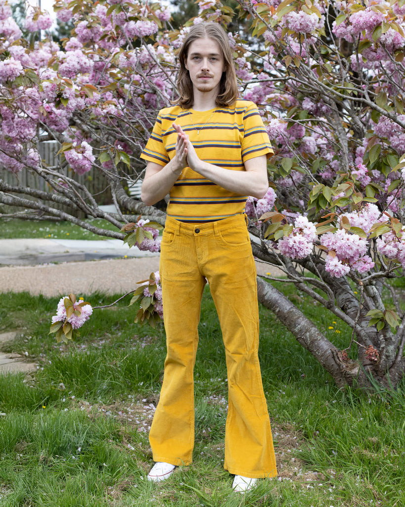  Model Jack stands in front of a cherry blossom tree wearing Run & Fly’s Gold Corduroy Flares paired with a yellow striped T-shirt. The bright colours create a bold and vibrant look against the floral background.