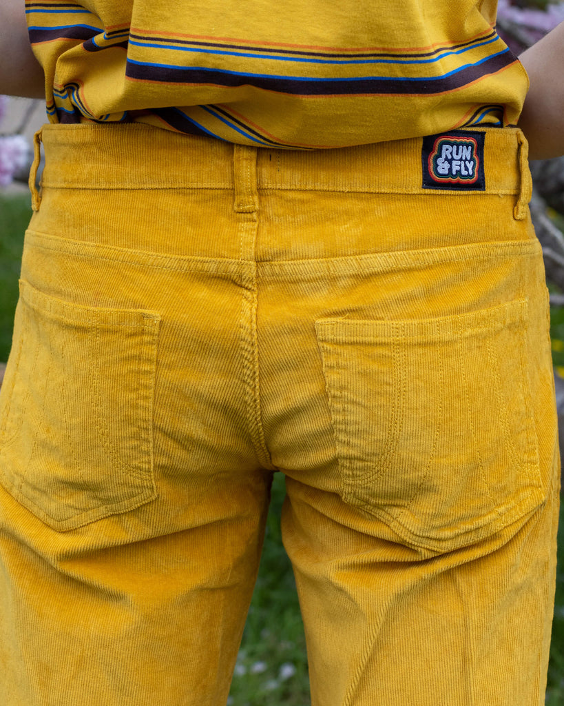Back view of model Jack wearing Run & Fly’s Gold Corduroy Flares, with the brand’s logo patch visible. The golden yellow fabric contrasts with the pink blossoms around him.