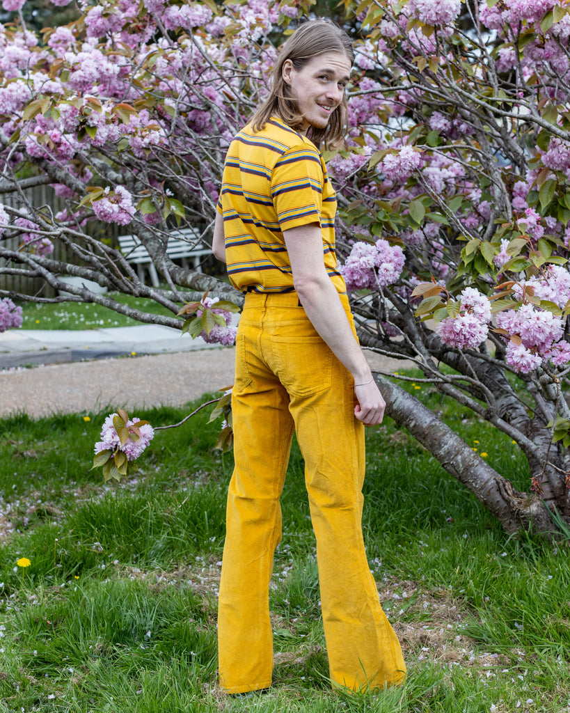 Model Jack stands outdoors in Run & Fly’s Gold Corduroy Flares, giving a playful over-the-shoulder look. He pairs the flares with a yellow striped T-shirt, blending harmoniously with the cherry blossom backdrop for a vibrant, retro-inspired style.