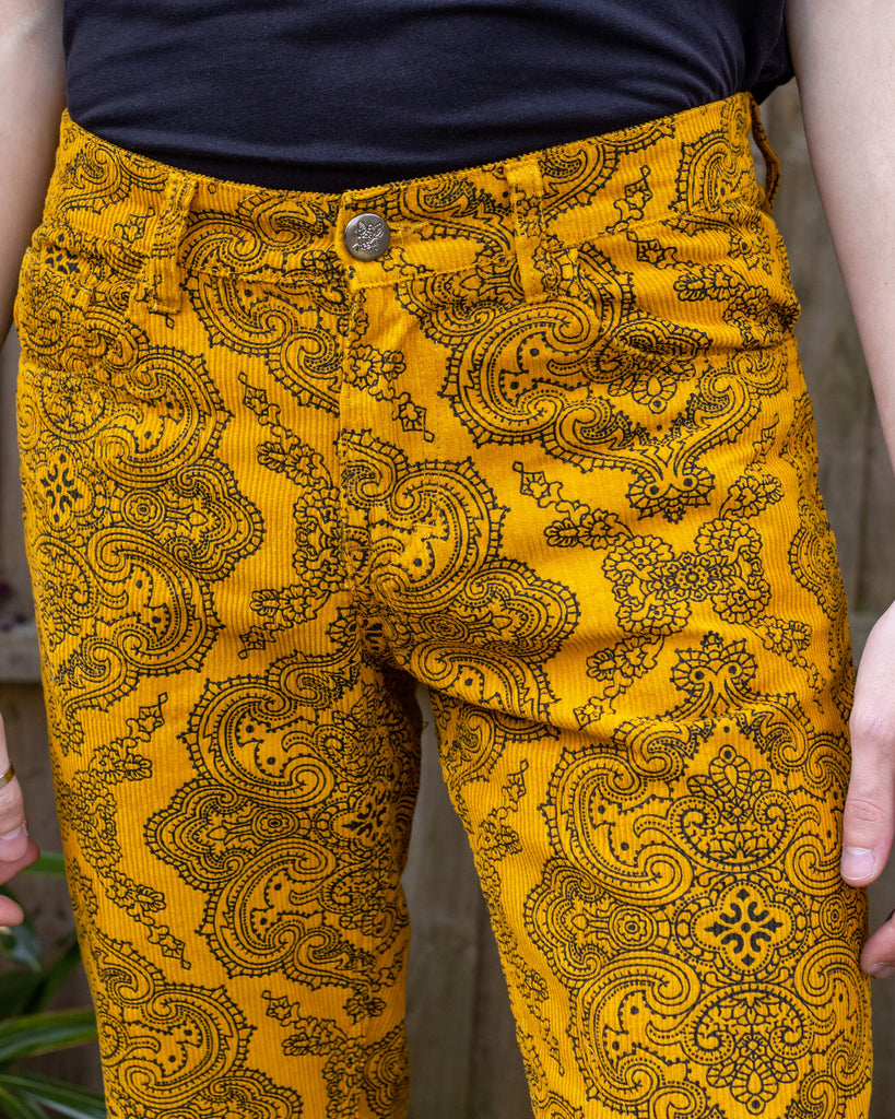 Close up of Jack wearing Gold Hendrix Paisley Corduroy Retro Bell Bottom Flares. The trousers are a yellow gold colour with an all over black paisley pattern. Close up shows the paisley pattern. The photo was taken outside in East Sussex.