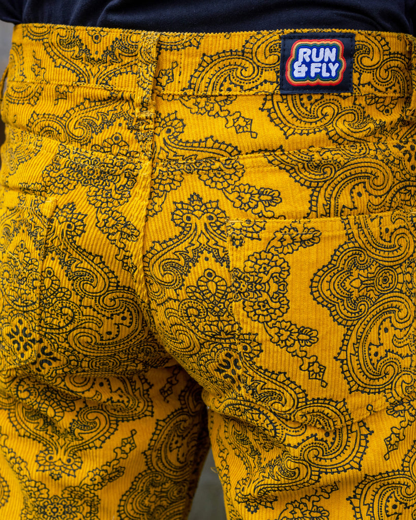 Close up of Jack wearing Gold Hendrix Paisley Corduroy Retro Bell Bottom Flares. The trousers are a yellow gold colour with an all over black paisley pattern. Close up shows the rainbow Run&Fly label on the back of the trousers.  The photo was taken outside in East Sussex.