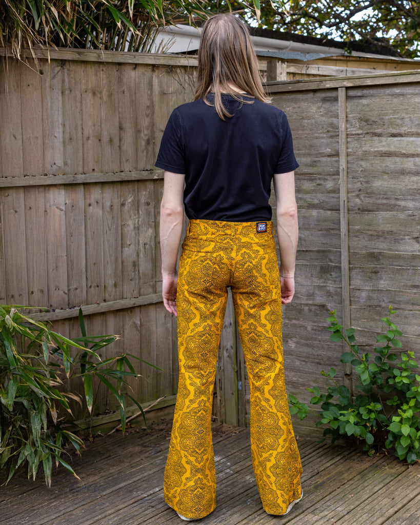 Jack, a white male model with long hair and glasses is wearing Gold Hendrix Paisley Corduroy Retro Bell Bottom Flares paired with a black t-shirt and brown shoes. The trousers are a yellow gold colour with an all over black paisley pattern. Jack is stood outside in front of a wooden fence with his back to the camera.