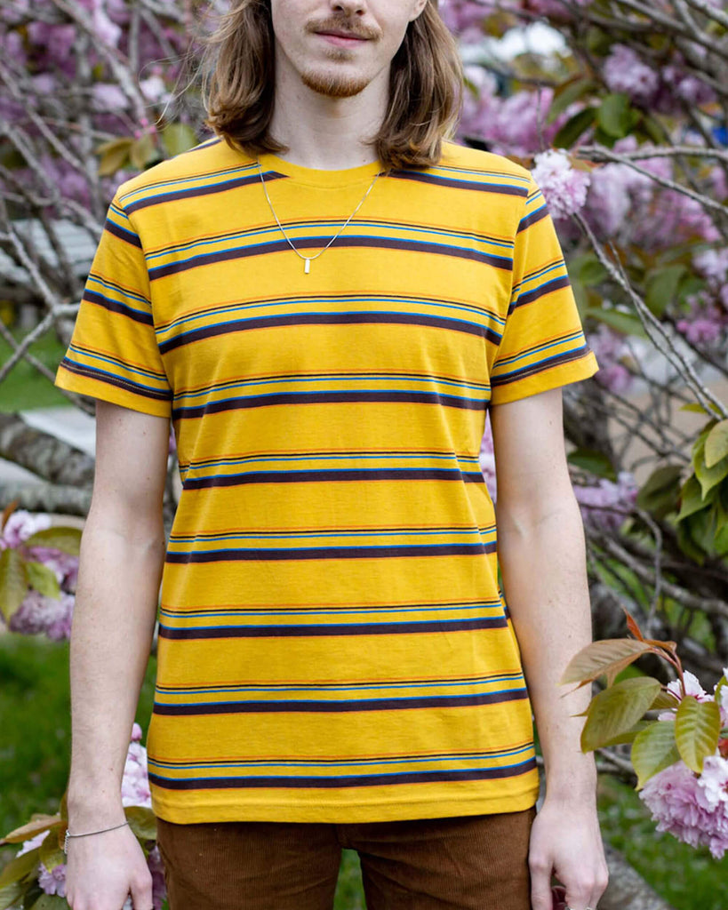 Jack is stood in front of a blossom tree wearing the gold sunset stripe retro tshirt with tobacco corduroy flares. He has mid length blonde hair and is posing facing towards the camera with both hands resting by his sides. Photo is cropped from the knees up.