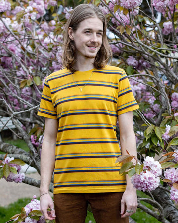 Jack is stood in front of a blossom tree wearing the gold sunset stripe retro tshirt with tobacco corduroy flares. He has mid length blonde hair and is smiling facing towards the camera with both hands resting by his sides. Photo is cropped from the knees up.