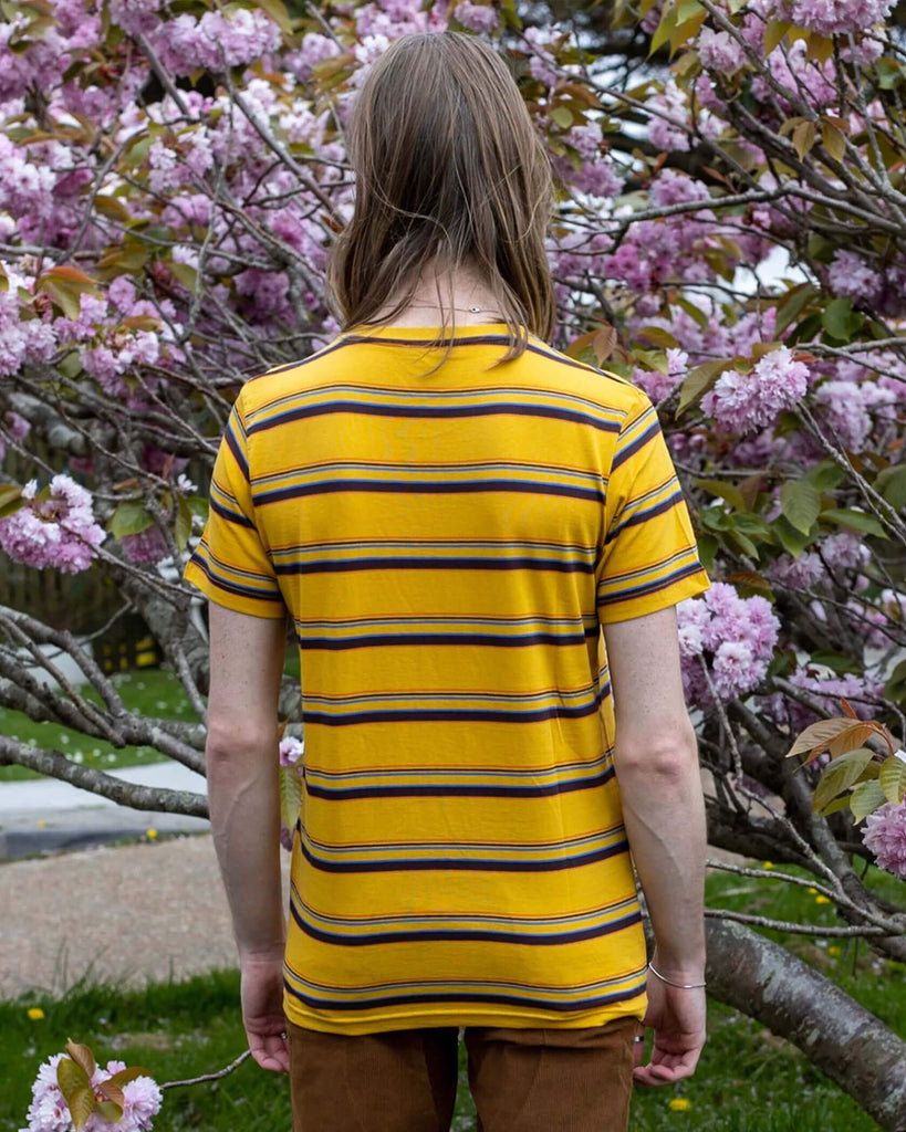 Jack is stood in front of a blossom tree wearing the gold sunset stripe retro tshirt with tobacco corduroy flares. He has mid length blonde hair and is posing facing away from the camera with both arms resting by his sides.